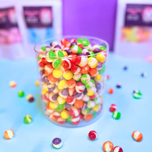 Gefriergetrocknete Rainbow Bites Fruits (vegan) fallen von oben in einen transparenten Glasbehälter – bunte Rainbow Bites landen sichtbar knusprig in der Mischung von Astrocat Candy.