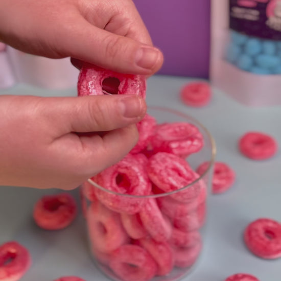 Ein gefriergetrockneter Erdbeerring aus Fruchtgummi wird in der Hand sichtbar knusprig gebrochen. Im Hintergrund steht ein Glasbehälter mit weiteren Erdbeerringen. Produkt von Astrocat Candy.