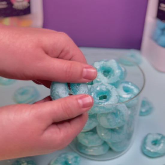 Ein knuspriger, gefriergetrockneter Blaubeerring wird von Hand in der Mitte gebrochen. Im Hintergrund ein Glasbehälter mit weiteren Blaubeerringen von Astrocat Candy.