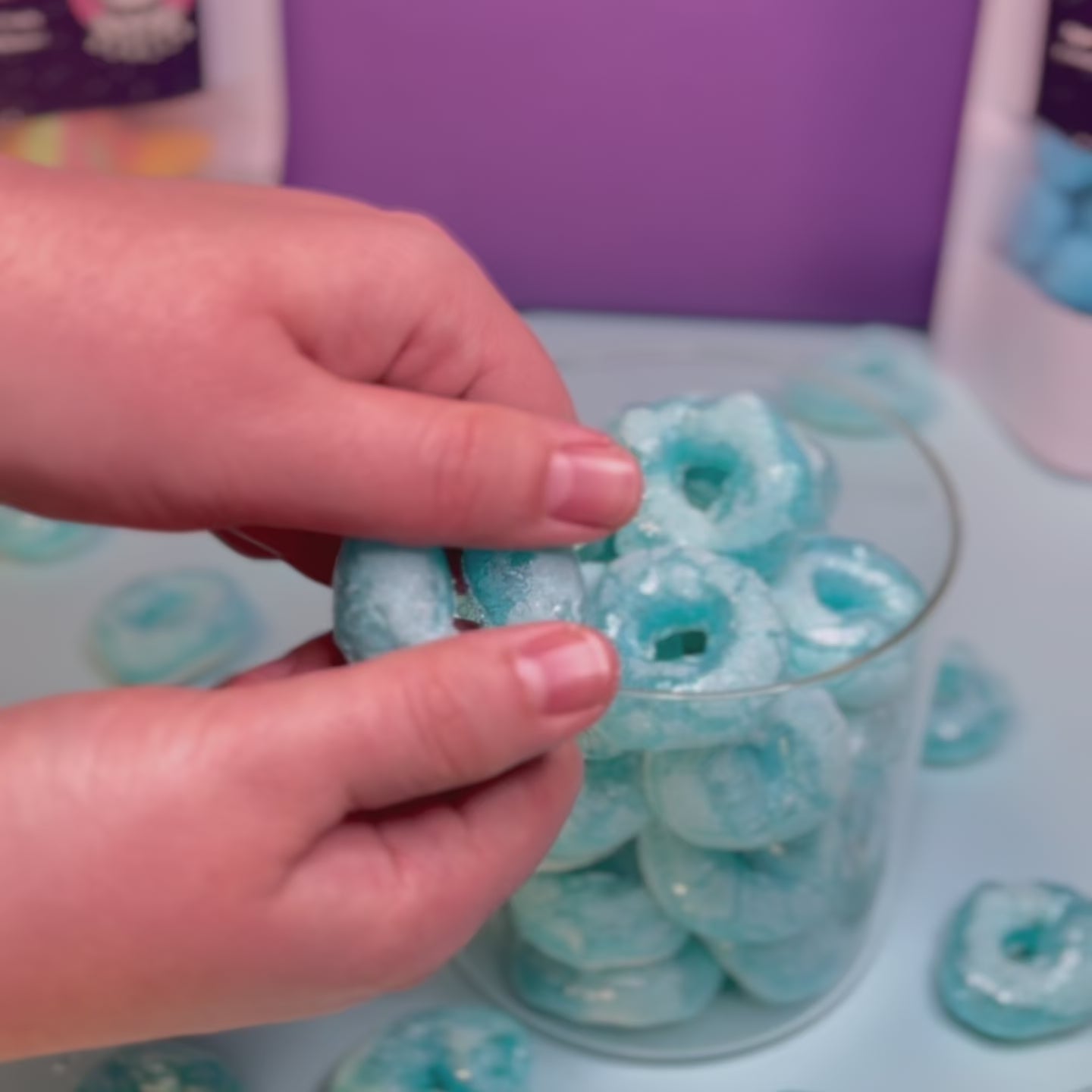 Ein knuspriger, gefriergetrockneter Blaubeerring wird von Hand in der Mitte gebrochen. Im Hintergrund ein Glasbehälter mit weiteren Blaubeerringen von Astrocat Candy.