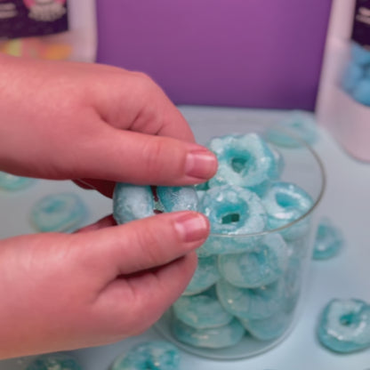 Ein knuspriger, gefriergetrockneter Blaubeerring wird von Hand in der Mitte gebrochen. Im Hintergrund ein Glasbehälter mit weiteren Blaubeerringen von Astrocat Candy.