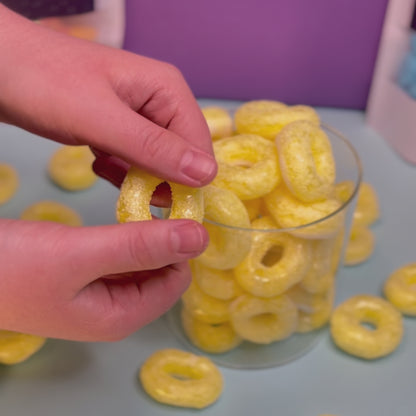 Ein gefriergetrockneter Bananenring wird in der Hand sichtbar knusprig gebrochen. Im Hintergrund steht ein Glas mit weiteren gefriergetrocknete Bananenringen von Astrocat Candy.
