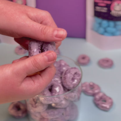 Ein gefriergetrockneter Passionsfruchtring wird von Hand geteilt – sichtbar knuspriger Bruch. Im Hintergrund ein Glas mit weiteren Ringen von Astrocat Candy