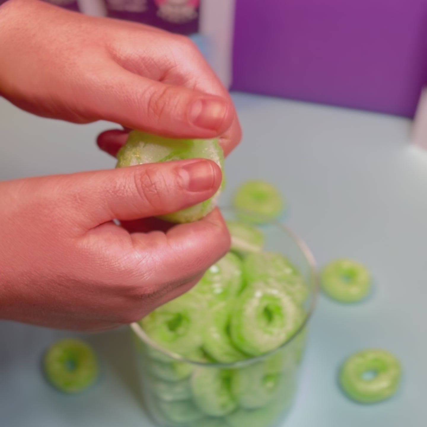 Ein gefriergetrockneter Apfelring (Halal) wird von Hand sichtbar knusprig in der Mitte gebrochen. Im Hintergrund steht ein Glasbehälter mit weiteren Apfelringen von Astrocat Candy.